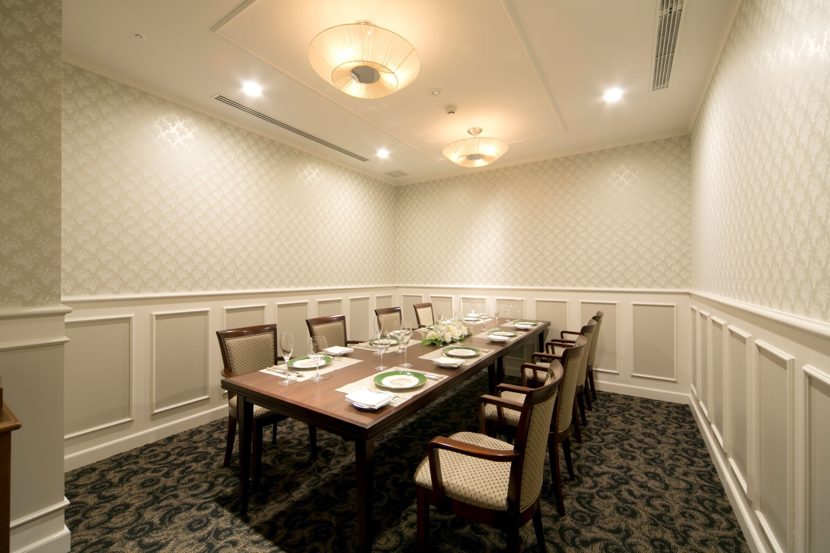A conference room with a long table set for a meeting, surrounded by chairs, with patterned wallpaper and recessed lighting.