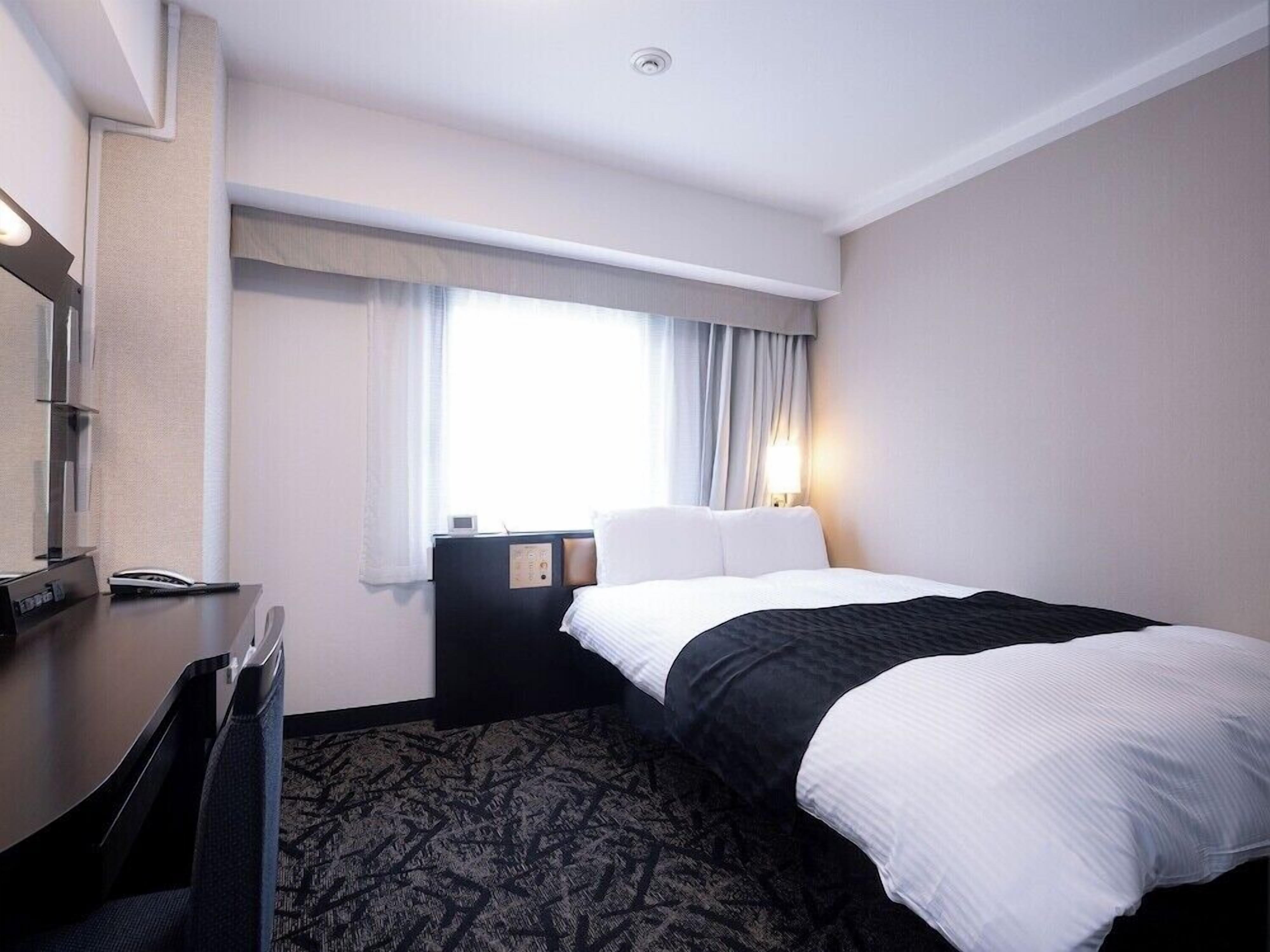 A hotel room with a bed, a desk, a chair, a nightstand, a lamp, and a window with curtains.