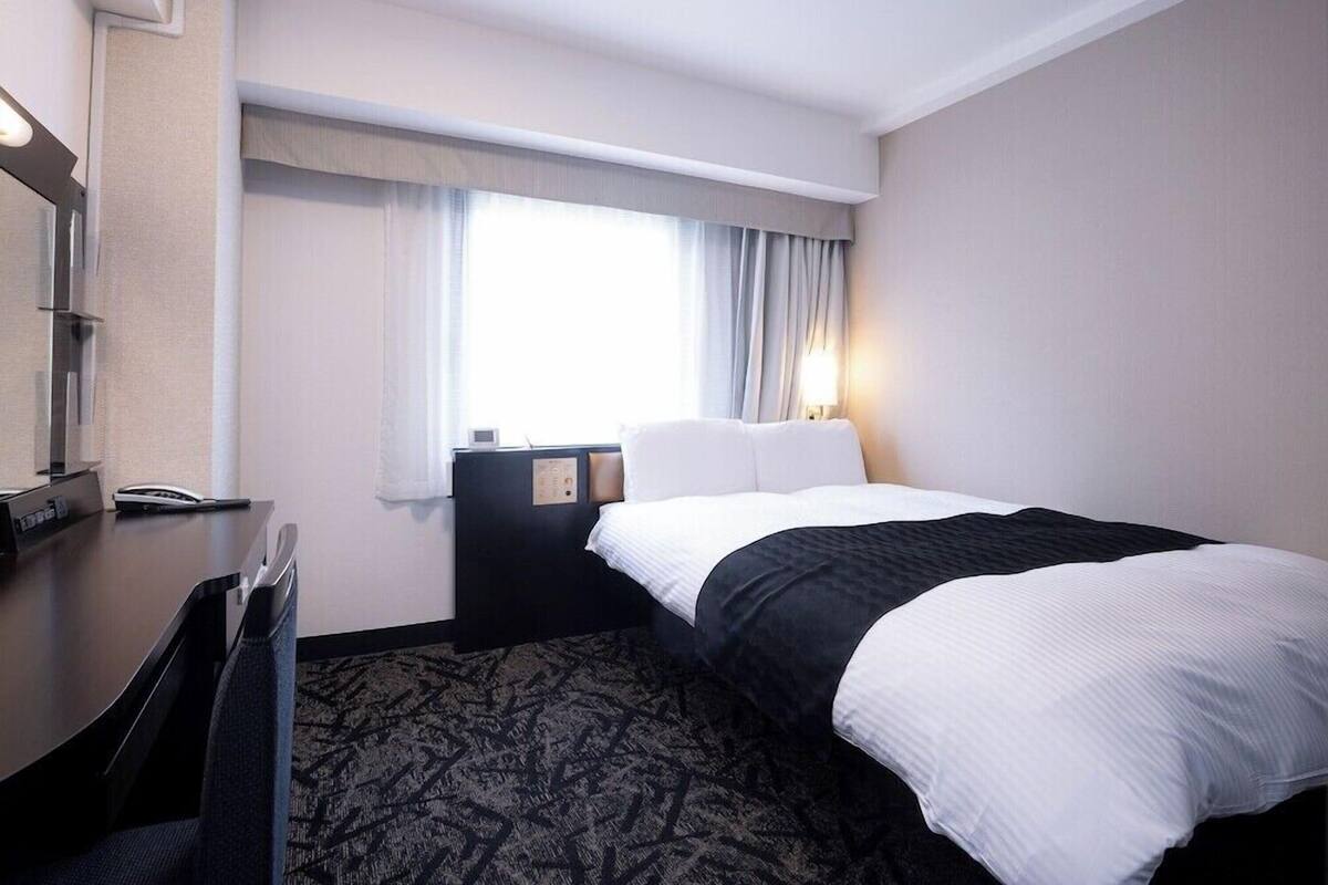 A hotel room with a bed, a desk, a chair, a nightstand, a lamp, and a window with curtains.