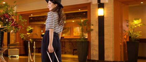 A woman in a lobby with a red suitcase, a striped shirt, and a black hat.