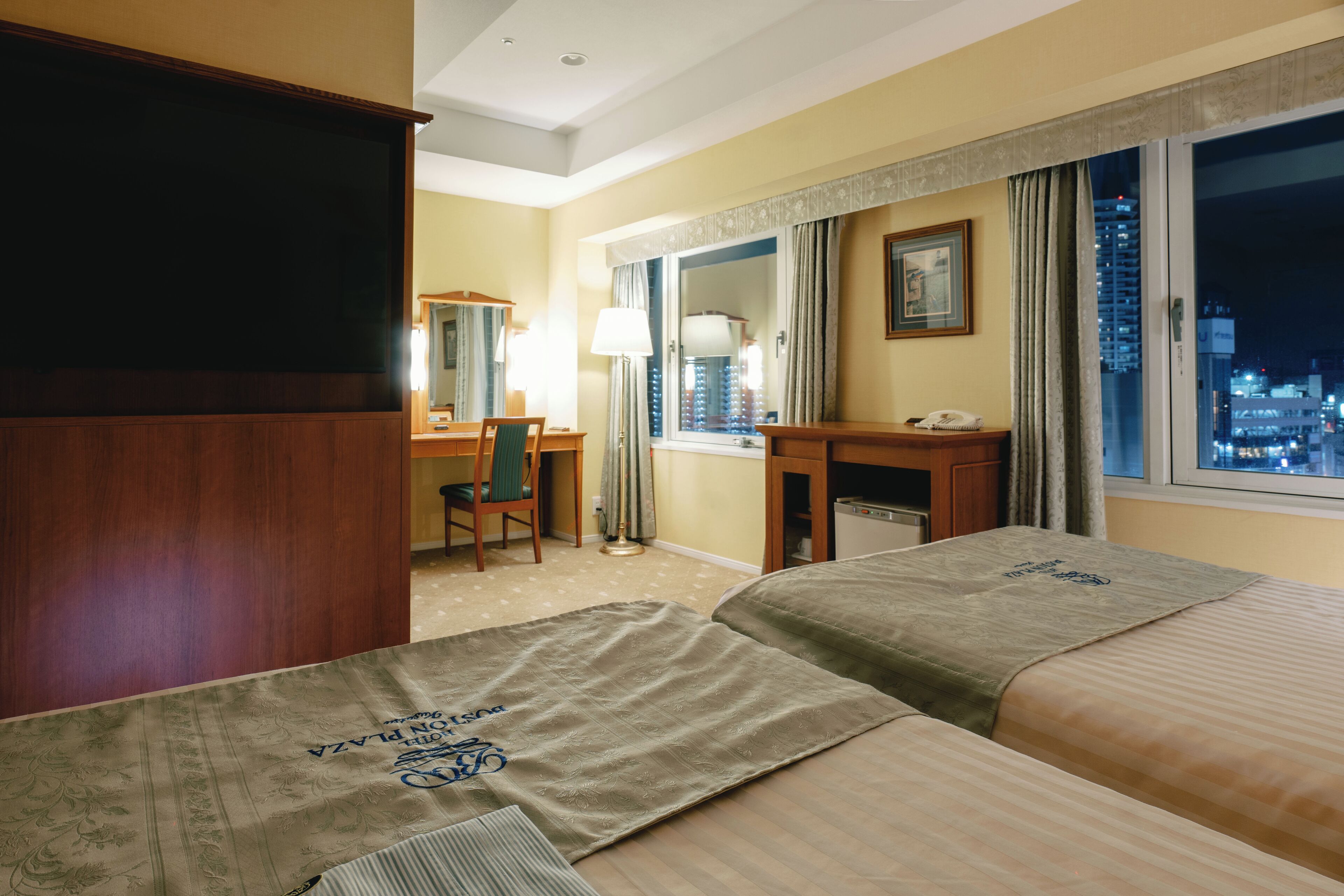 A hotel room with two beds, a desk, a chair, a lamp, a window with a city view, and a mirror.