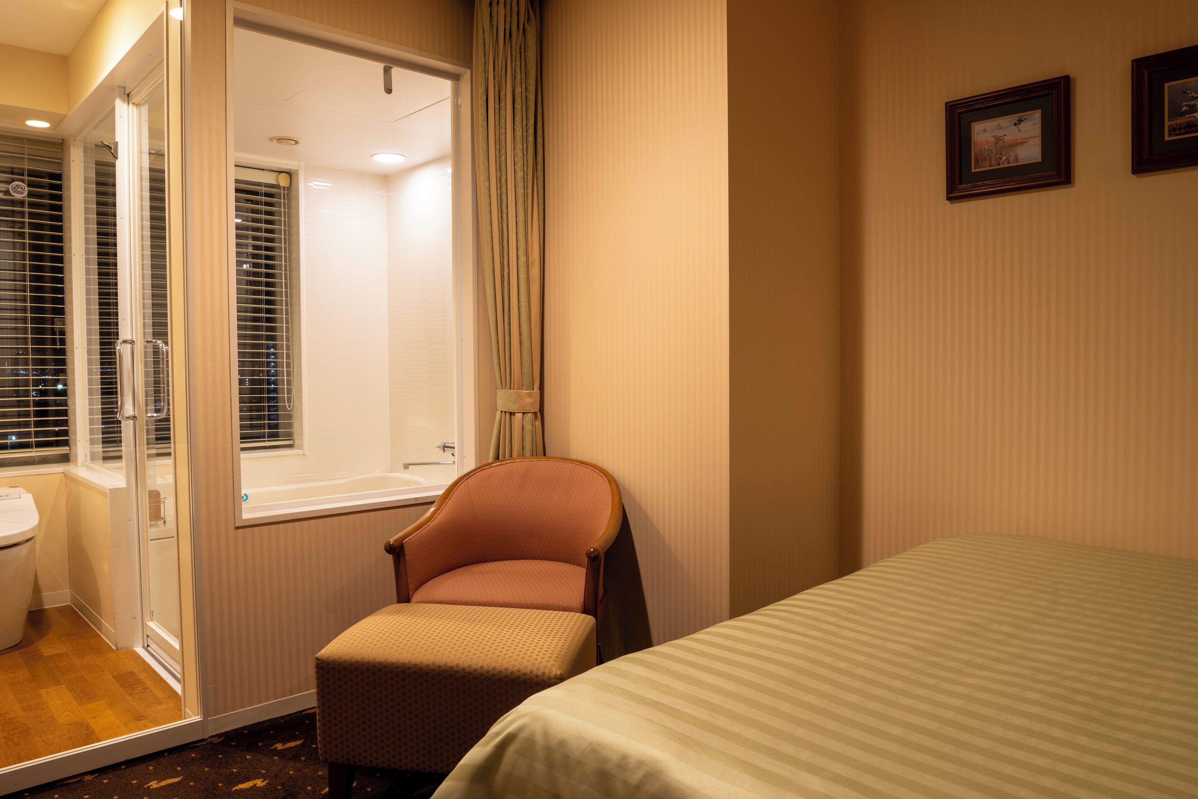 Una camera d'albergo con un letto, una sedia, un bagno con il water e una finestra con tende.