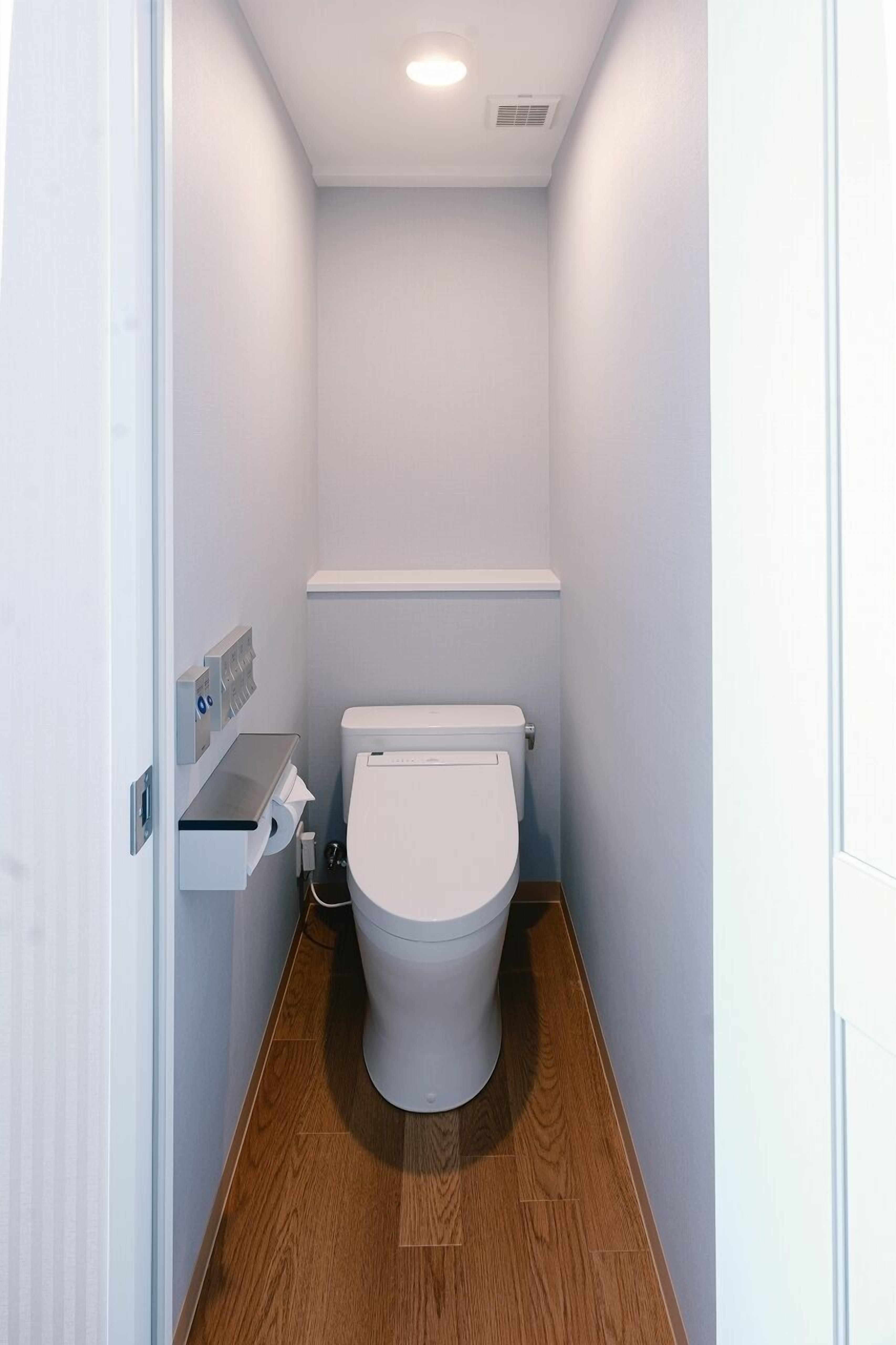 Un baño estrecho con paredes blancas, un inodoro blanco, suelo de madera y un estante pequeño con dispensador de toallas de papel.