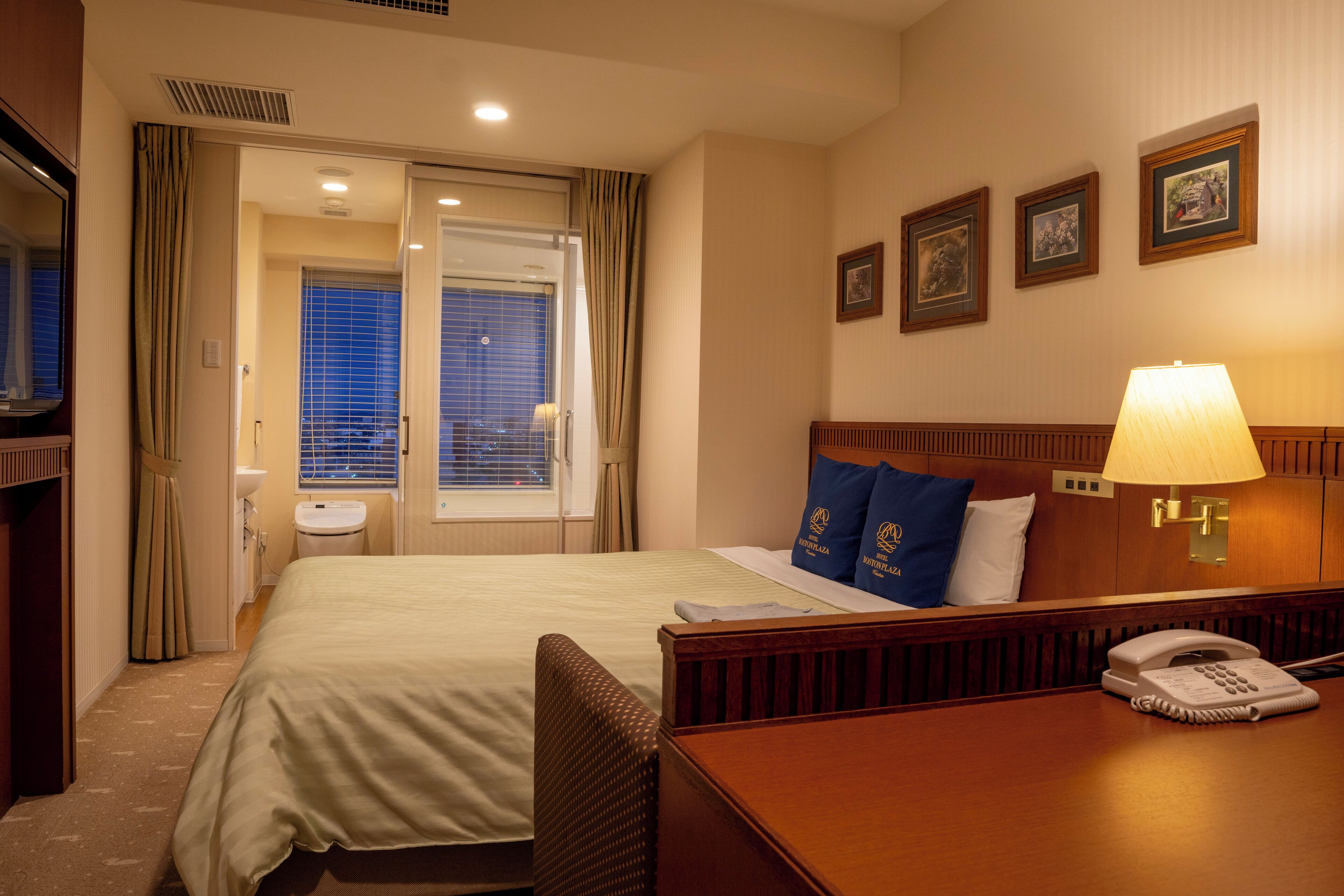 Una habitación de hotel con cama, mesita de noche con lámpara, teléfono y un muro decorado con marcos.