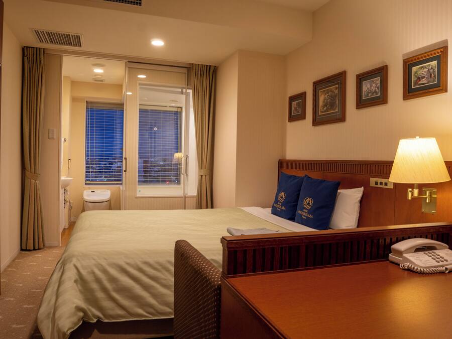Una habitación de hotel con cama, mesita de noche con lámpara, teléfono y un muro decorado con marcos.