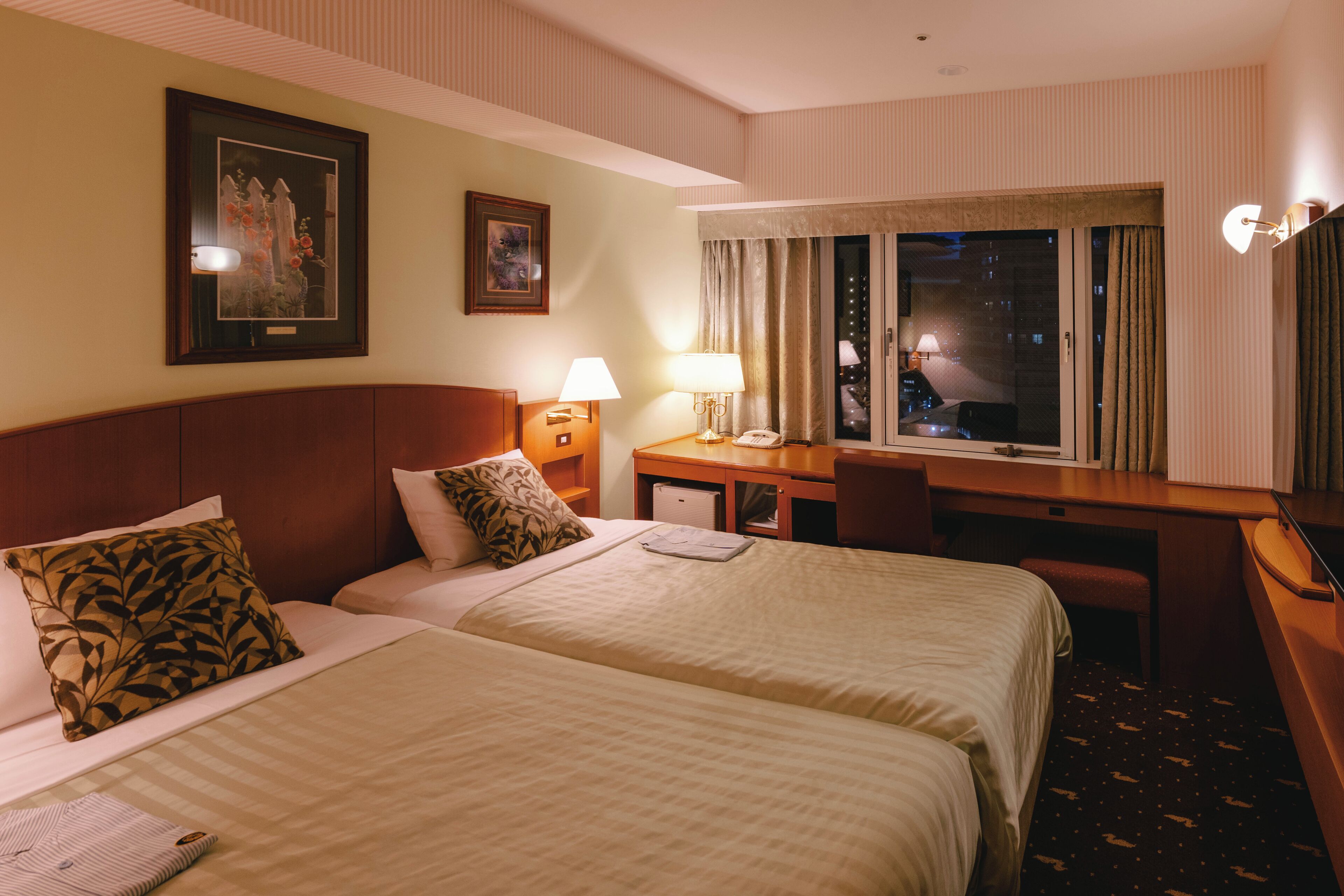 A hotel room with two beds, a desk, a chair, a desk lamp, a telephone, and a window with curtains.