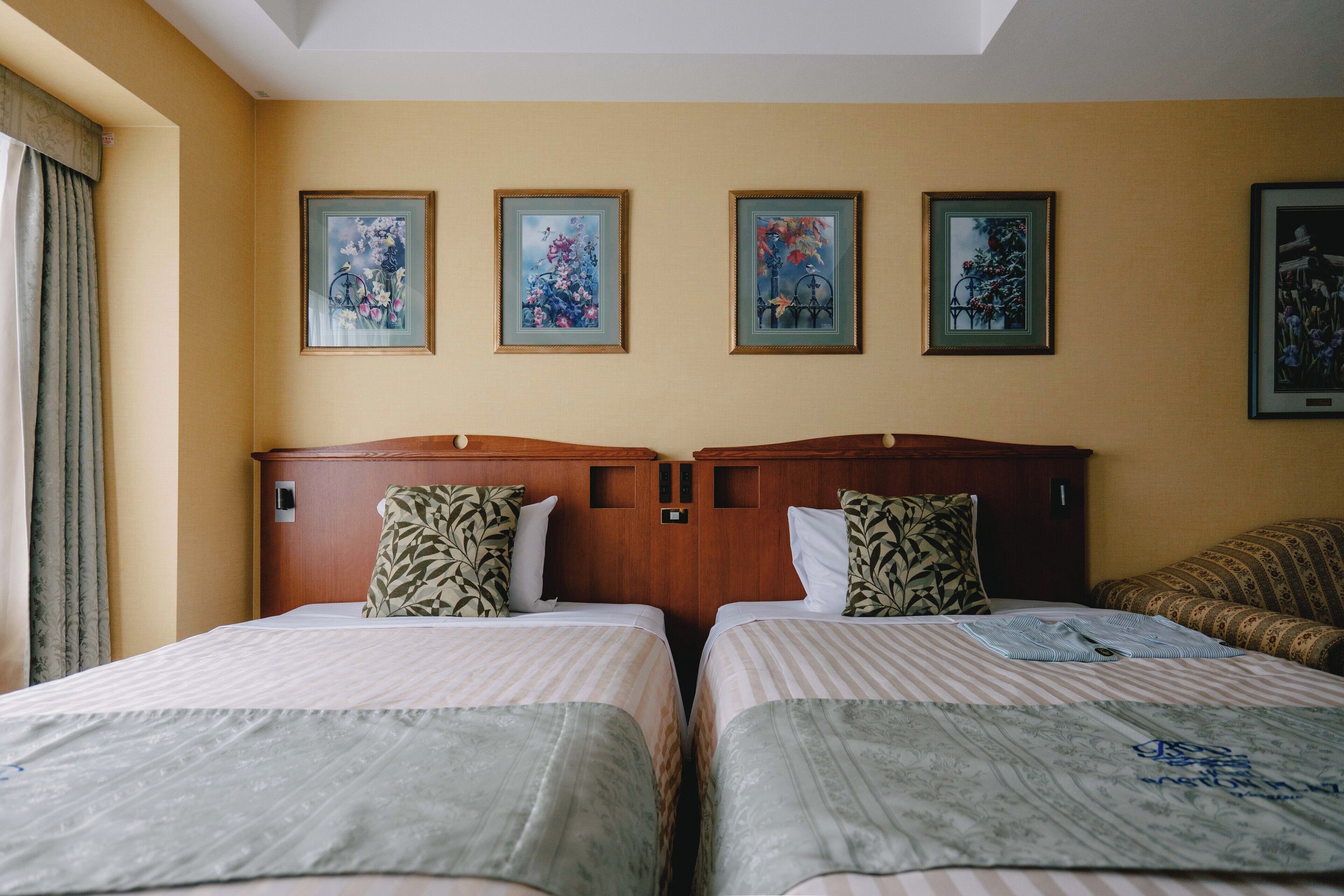 A hotel room with two beds, a wooden headboard, and four framed floral paintings on the wall.