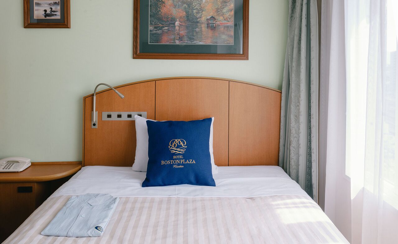 Een hotelkamer met een houten hoofdbord, een blauw kussen met het logo van het hotel, een bedtelefoon en een raam met gordijnen.