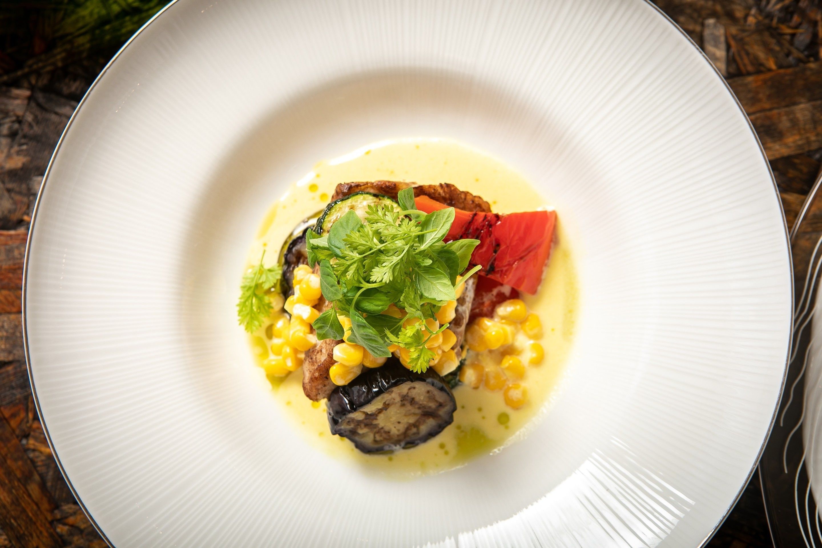 En hvid tallerken med en ret bestående af majs, aubergine og grønt, serveret på et træbord.