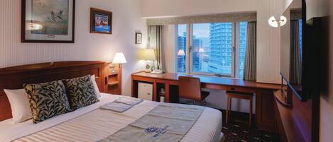 A hotel room with a large bed, a desk with a chair, a television, and a window with a view of buildings.