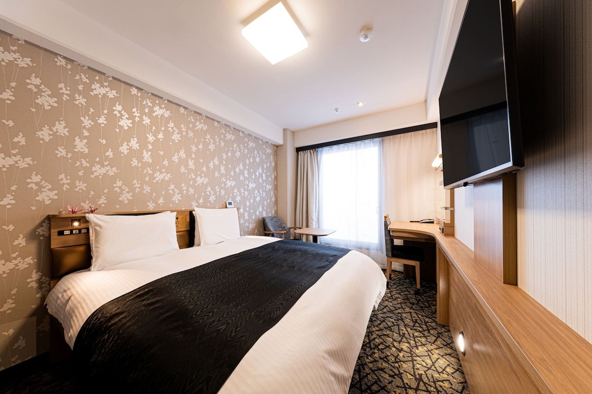 Una habitación de hotel con cama, escritorio, televisión y ventana con cortinas.