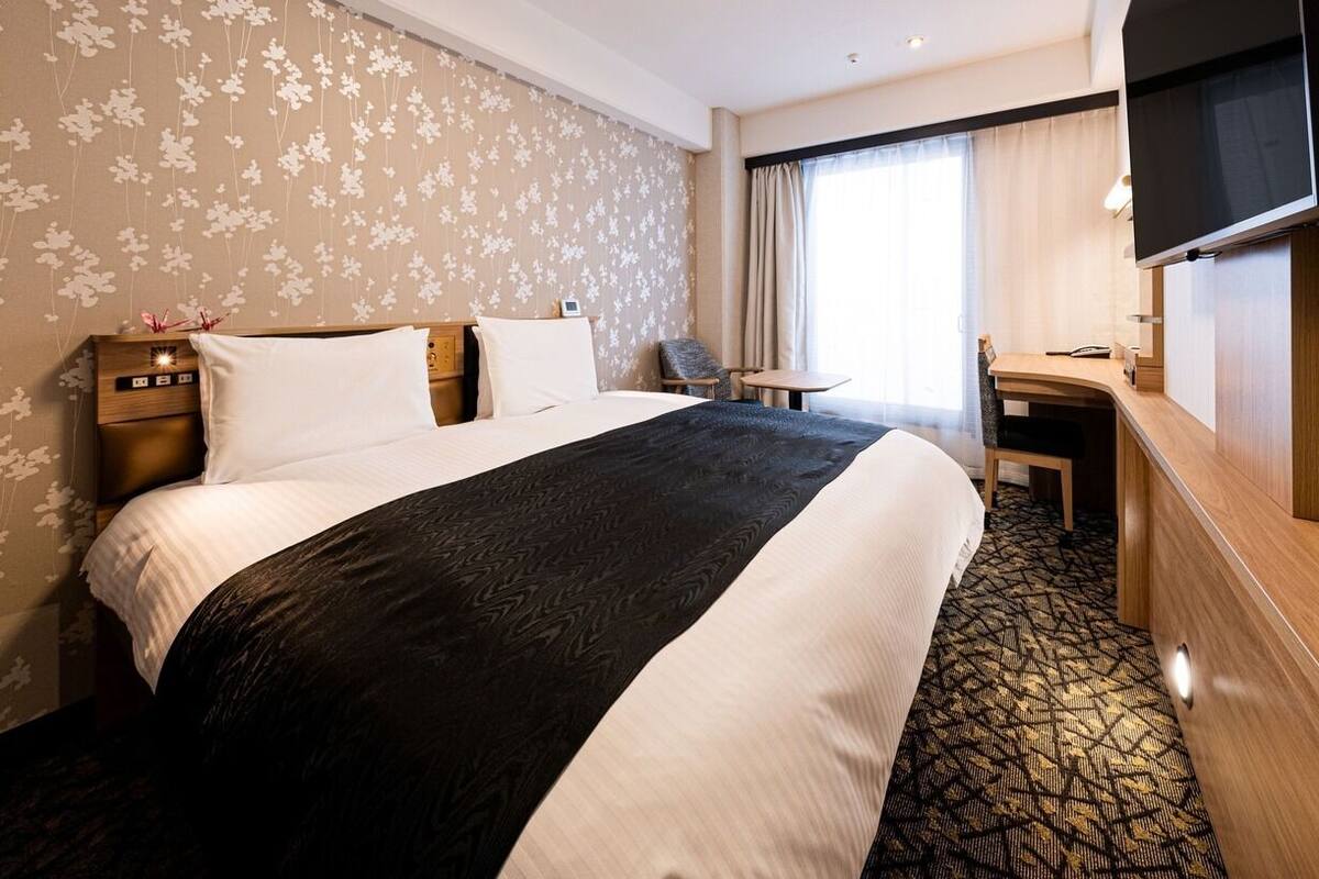 Una habitación de hotel con cama, escritorio, silla, televisor y ventana con cortinas.