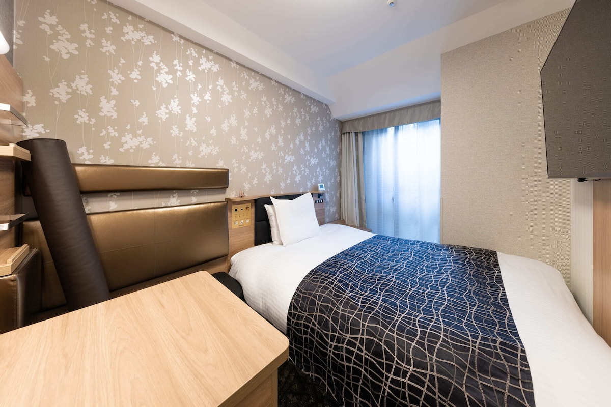 Habitación de hotel con cama, escritorio, silla, panel de control y ventana con cortinas.