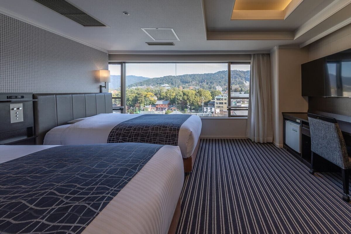 Quarto de hotel com duas camas, uma mesa de trabalho, televisão e uma grande janela com vista para a cidade e montanhas.