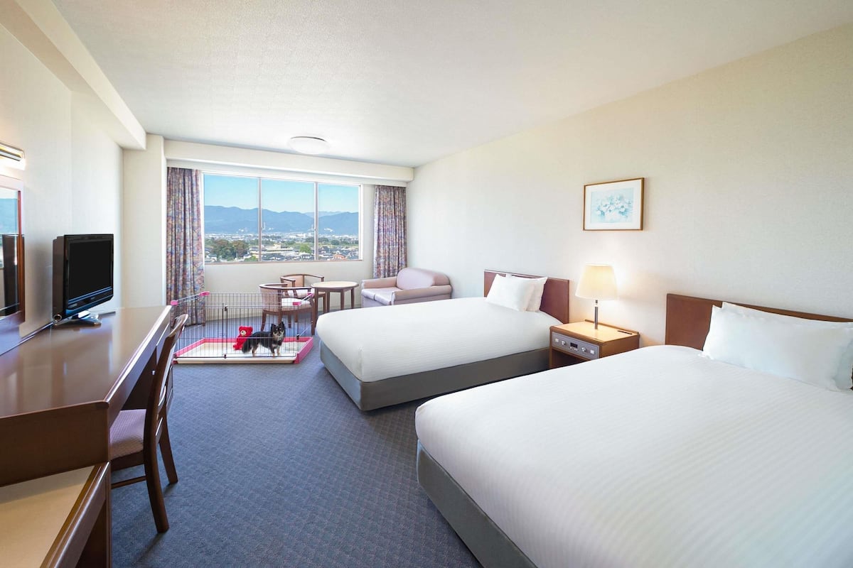 A hotel room with two beds, a desk, a TV, a chair, a small table, a lamp, and a view of the city and mountains.