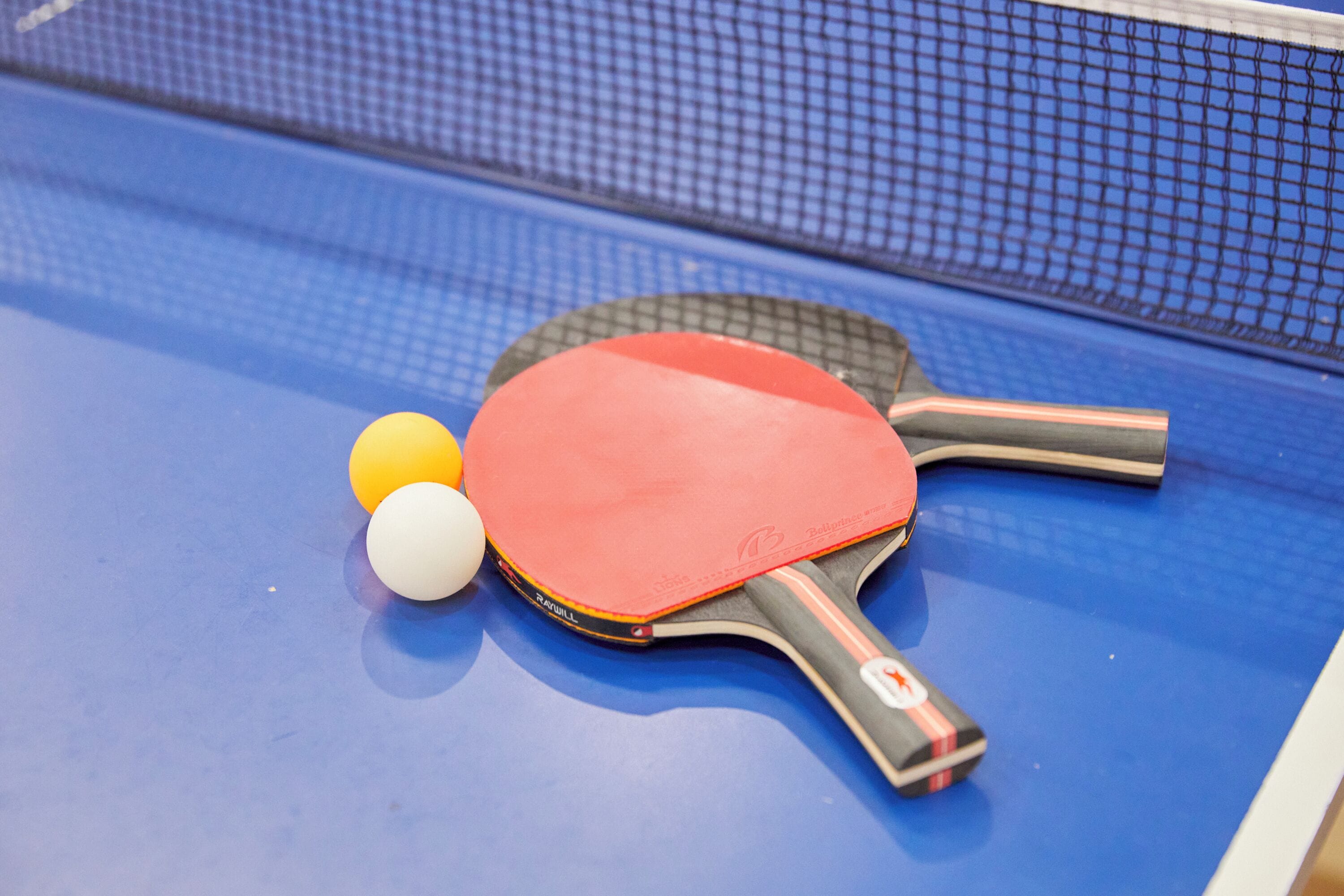 A table tennis table with a blue surface, a red table tennis paddle, and two table tennis balls.