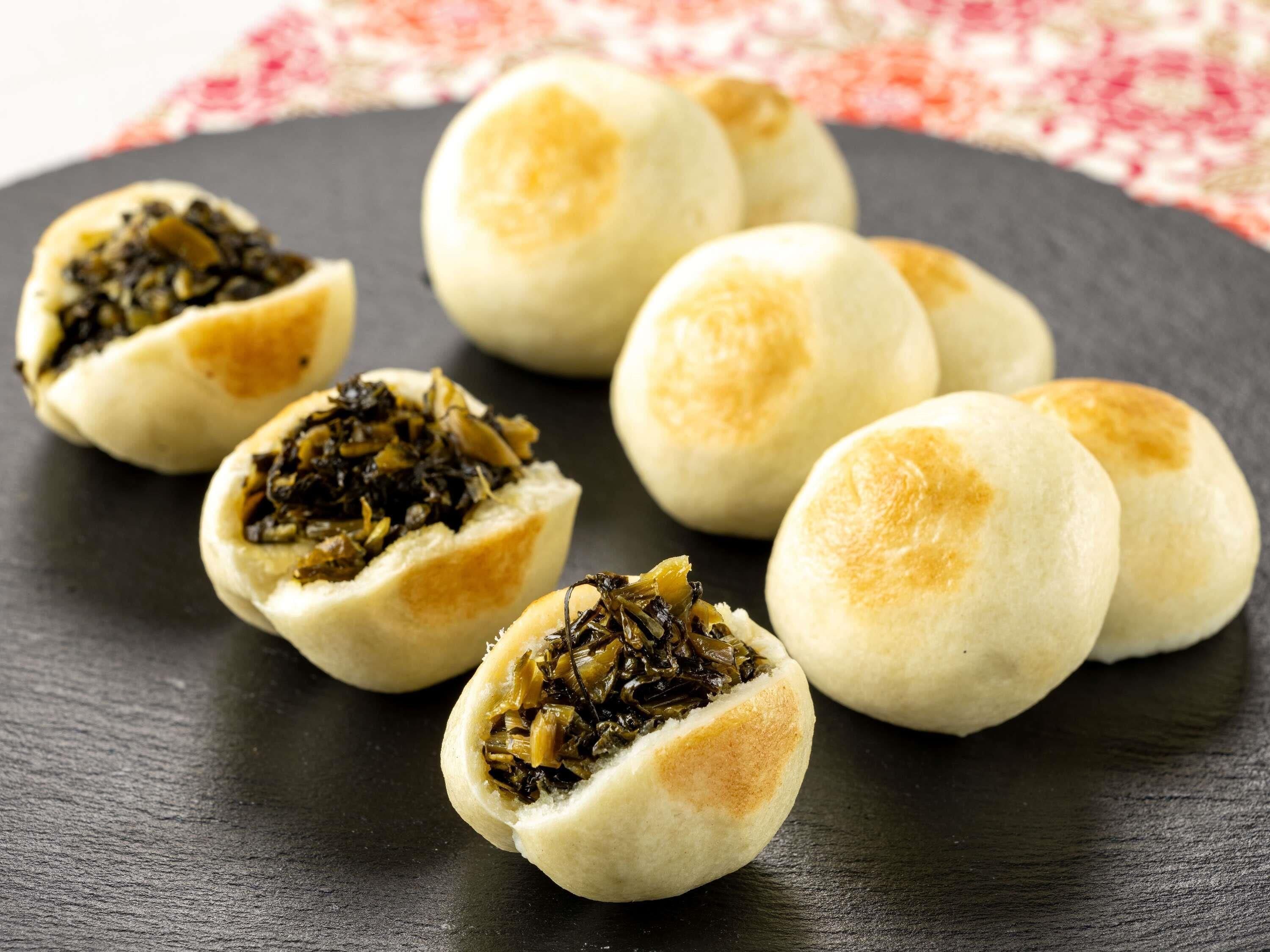 Ein schwarzes Schiefertablett mit mehreren runden, goldbraunen Brötchen, von denen einige aufgeschnitten eine Füllung aus dunkelgrünem Gemüse zeigen.