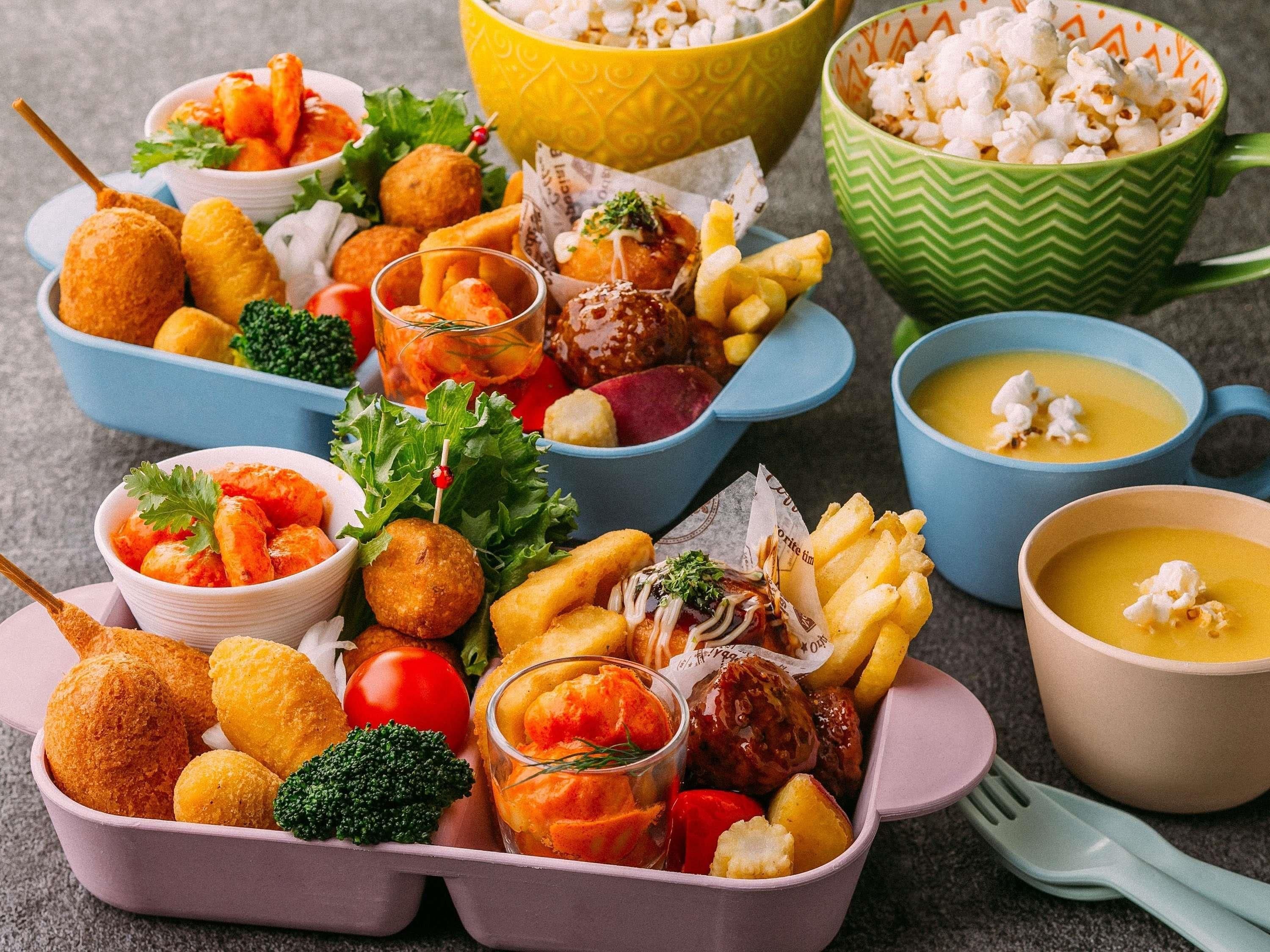 Diverse soorten eten, waaronder gefrituurde snacks, groenten en soep, geserveerd in kommen en bakjes in verschillende kleuren.