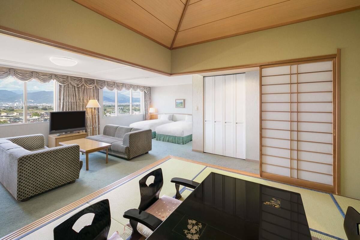 A hotel room with a large window, a bed, a TV, a dining table, and a sliding door.