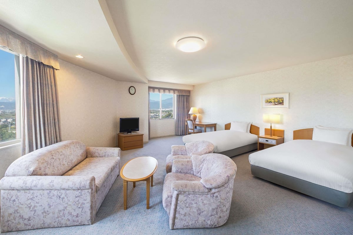 A hotel room with a large bed, two sofas, a small table, a TV on a stand, a desk with a lamp, and a window with a view of the city.