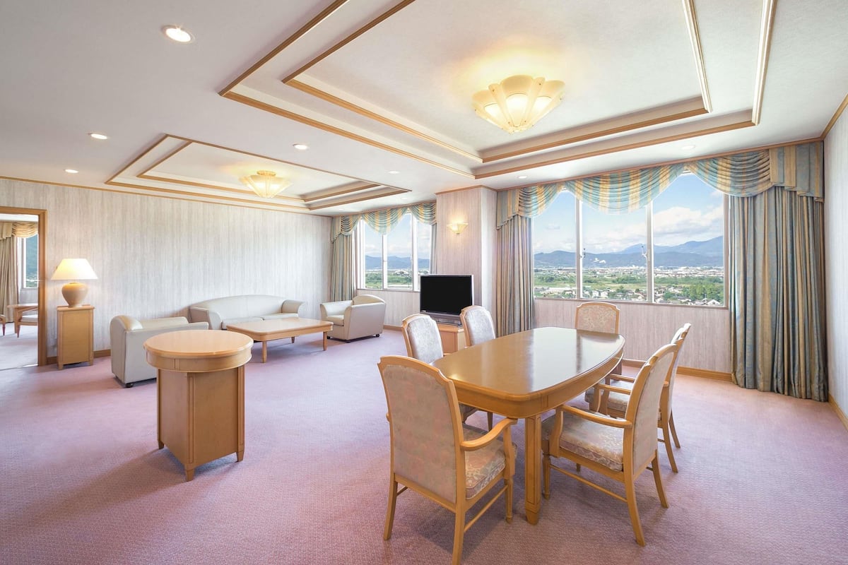 Ein Hotelzimmer mit großem Esstisch, bequemen Sitzgelegenheiten, einem Fernseher und Blick auf die Landschaft.