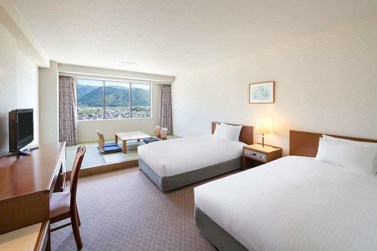Ein Hotelzimmer mit zwei Betten, einem Schreibtisch mit Fernseher, einem Sessel, einem Tisch und Blick auf Berge.