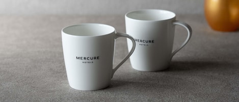 Deux tasses blanches ornées de l’inscription « MERCURE HOTELS », posées sur une surface texturée avec un arrière-plan flou.