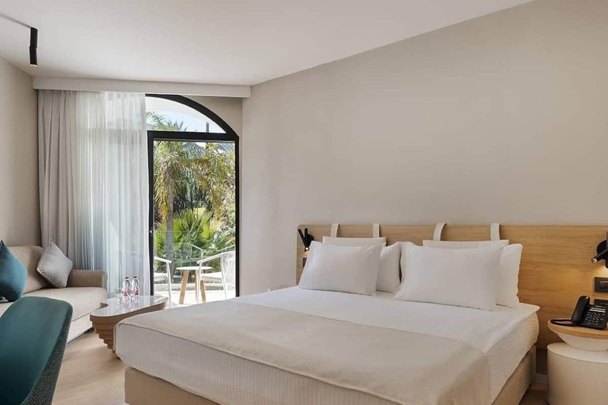 Une chambre d’hôtel moderne, dotée d’un grand lit, d’un canapé, d’un fauteuil et d’une petite table. Elle dispose également d’un balcon avec vue sur la verdure.