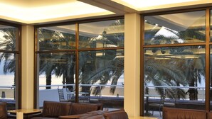 Un salon moderne doté de canapés et de fauteuils en cuir, de grandes fenêtres offrant une vue sur des palmiers et une plage, et d’un plafond pourvu d’un éclairage encastré.