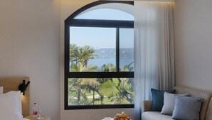 Une chambre d’hôtel avec un lit, un canapé, une petite table avec des fruits, une fenêtre donnant sur des palmiers et un plan d’eau, et un luminaire au plafond.