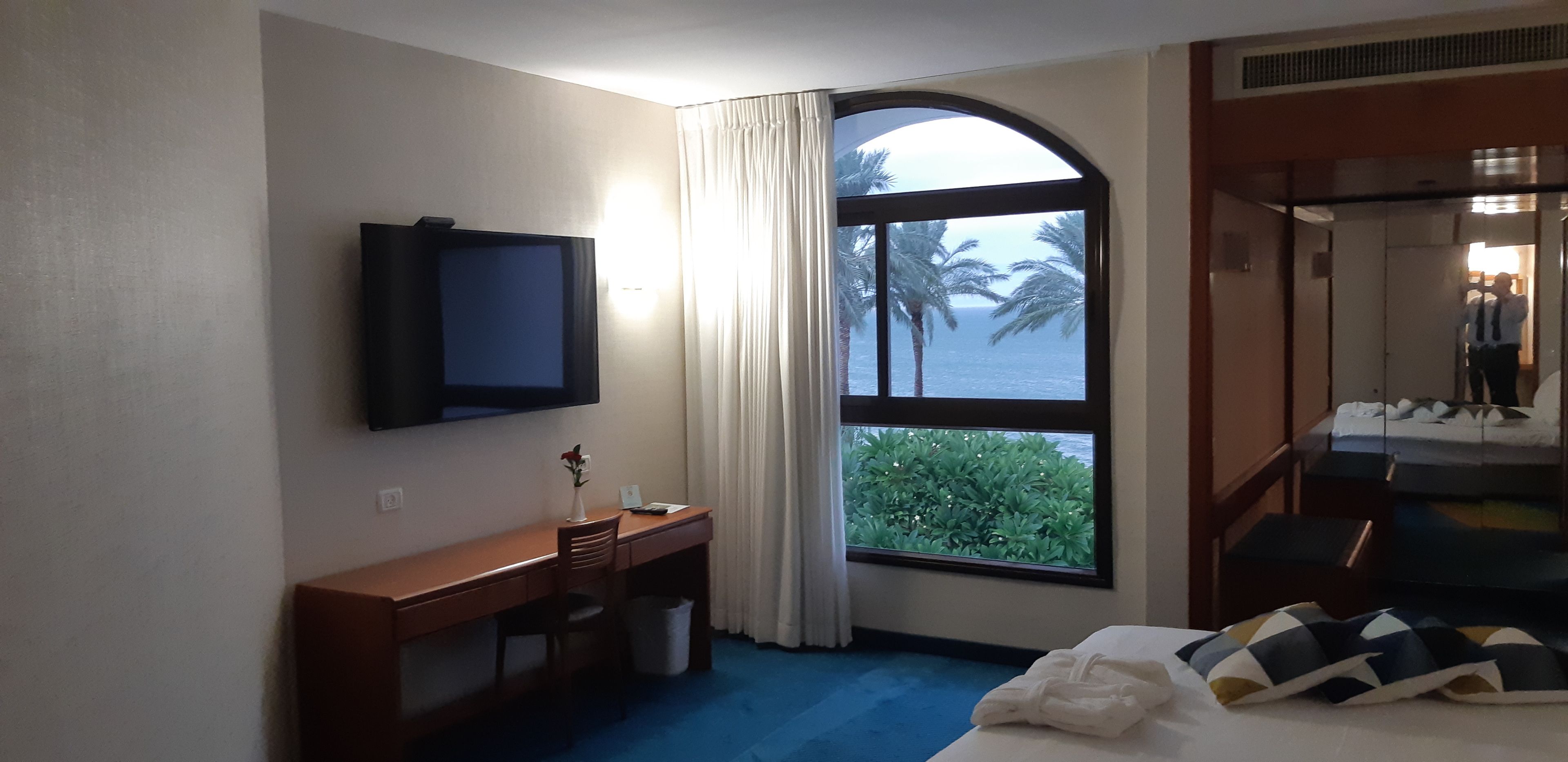 Une chambre d’hôtel comprenant un lit, un bureau avec une chaise, une télévision, une grande fenêtre donnant sur des palmiers et la mer, et un miroir.