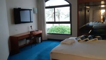 Habitación de hotel con cama, escritorio, silla, televisión y ventana con vista a palmeras.