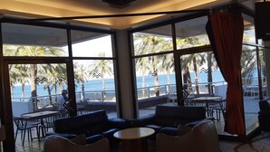 Un vaste espace intérieur doté de grandes fenêtres offrant une vue sur la mer et les palmiers. On y trouve plusieurs coins salon avec des tables et des chaises.