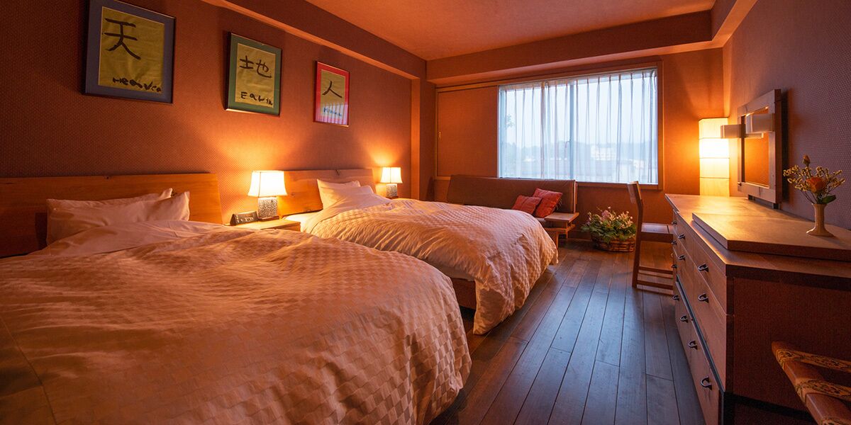 A hotel room with two beds, bedside lamps, a desk with a chair, and a window with curtains.