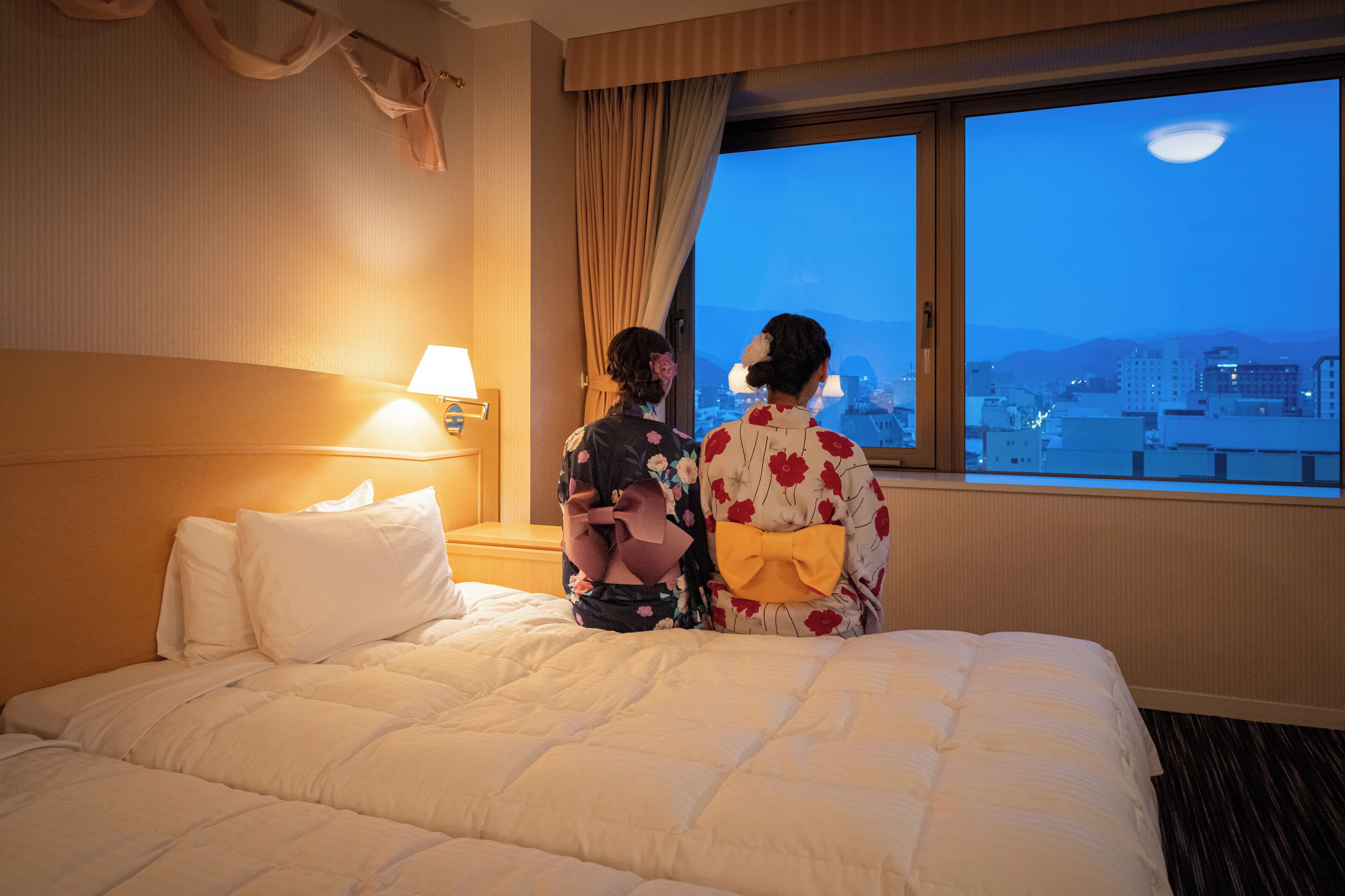Habitació d'hotel amb un llit gran, una taula de nit amb una làmpada i vista a la ciutat a través d'una finestra.
