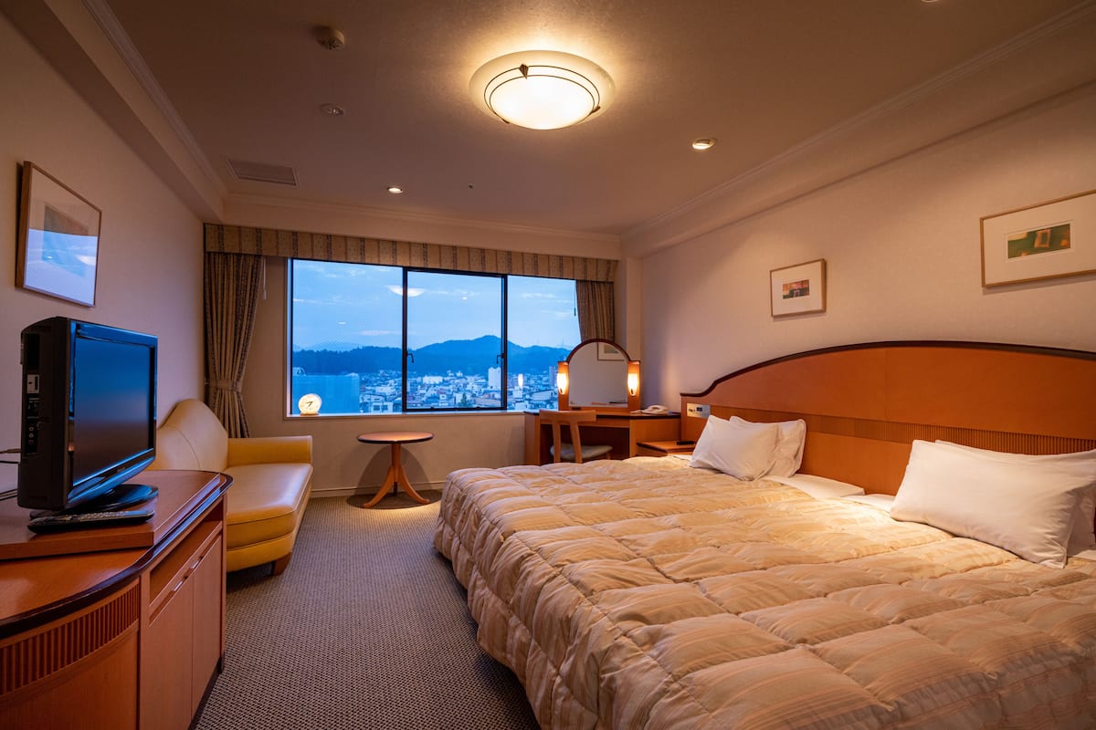 Habitación de hotel con una cama grande, un escritorio, un televisor y vistas a un paisaje urbano desde la ventana.