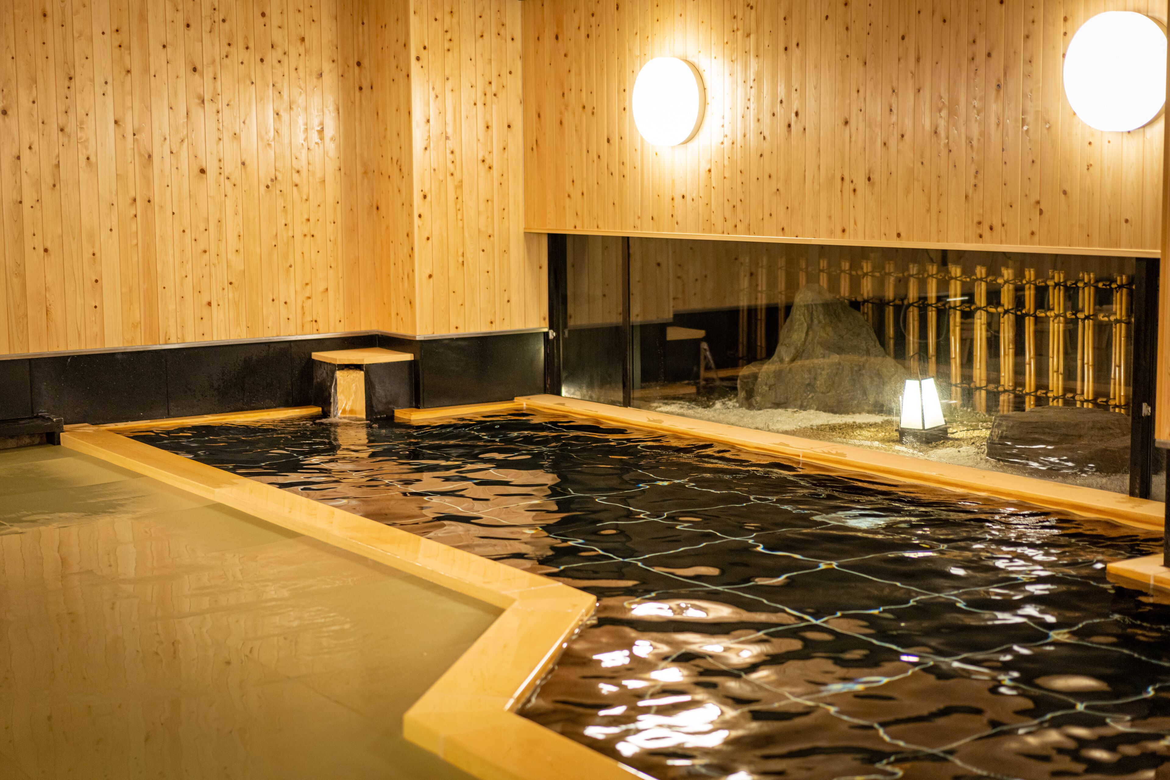 A wooden interior with a stone-tiled hot spring bath, a small wooden stool, and a bamboo shelf.