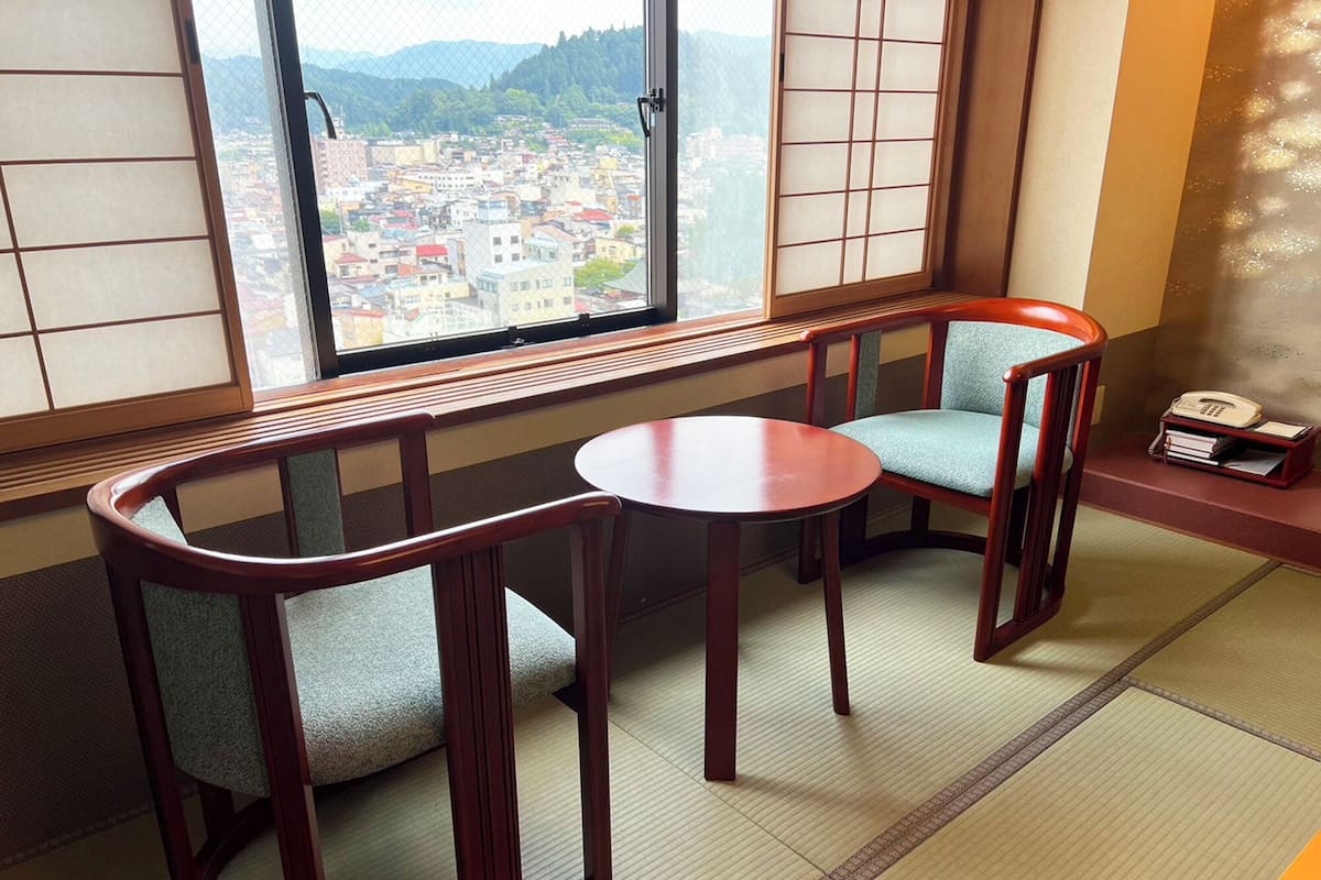 Una habitación japonesa tradicional con suelo de tatami, una mesa baja, dos sillas acolchadas y vistas a un pueblo a través de un shoji.