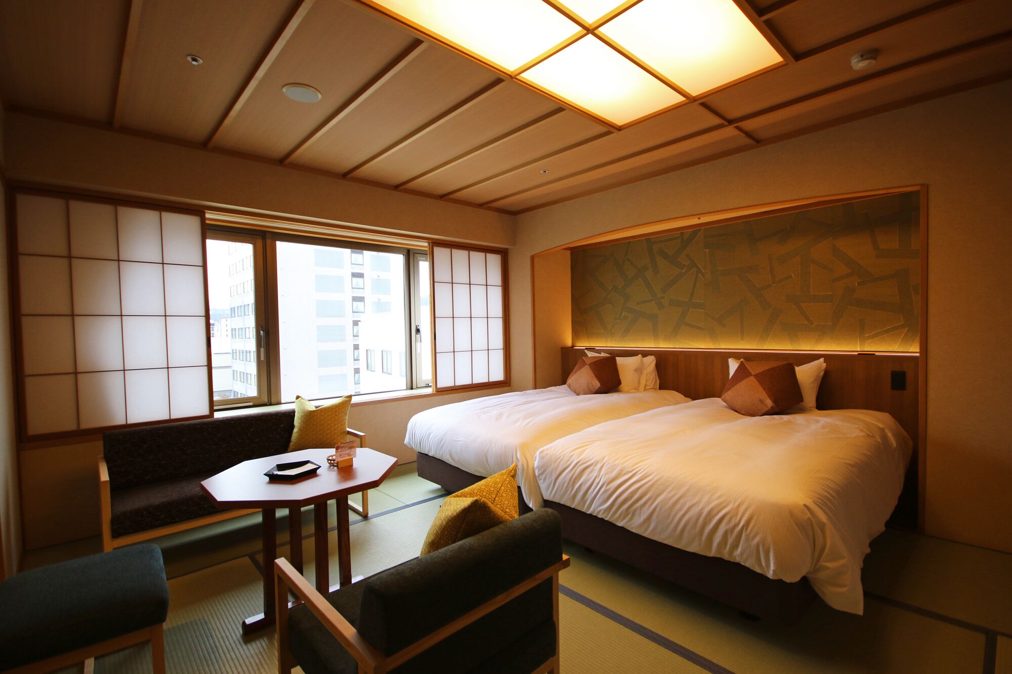 Quarto de hotel com uma cama grande, uma mesinha, um sofá e vista para edifícios pela janela.