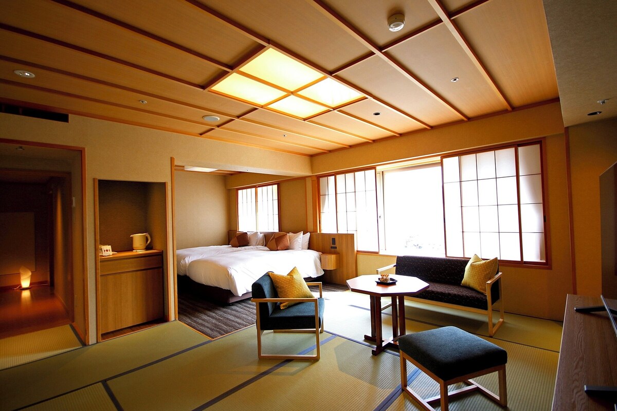 Ein traditionelles Hotelzimmer im japanischen Stil mit Tatami-Böden, einem Bett, einer Couch, einem kleinen Tisch und einem Schreibtisch.