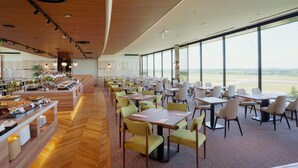A spacious dining area with multiple tables and chairs, a buffet counter with food items, and large windows offering a view of the outdoors.