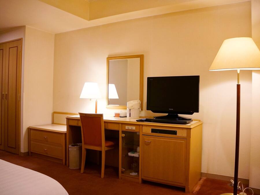 A hotel room with a bed, a desk with a chair, a television, a lamp, and a wardrobe.