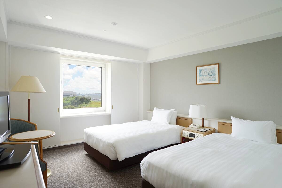 A hotel room with two beds, a desk, a chair, a TV, a window with a view, and a painting on the wall.