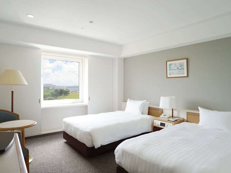 Quarto de hotel com duas camas, uma escrivaninha, uma cadeira, televisão, janela com vista e um quadro na parede.