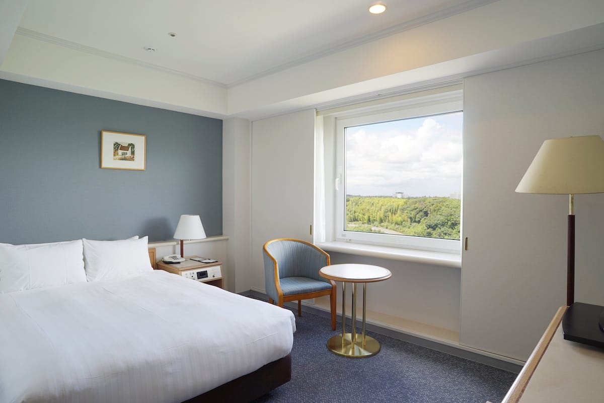 A hotel room with a bed, a chair, a small table, a lamp, a nightstand, and a window with a view.