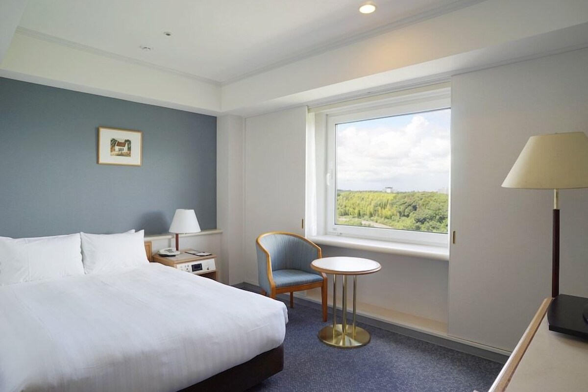 A hotel room with a bed, a chair, a small table, a lamp, and a window with a view.