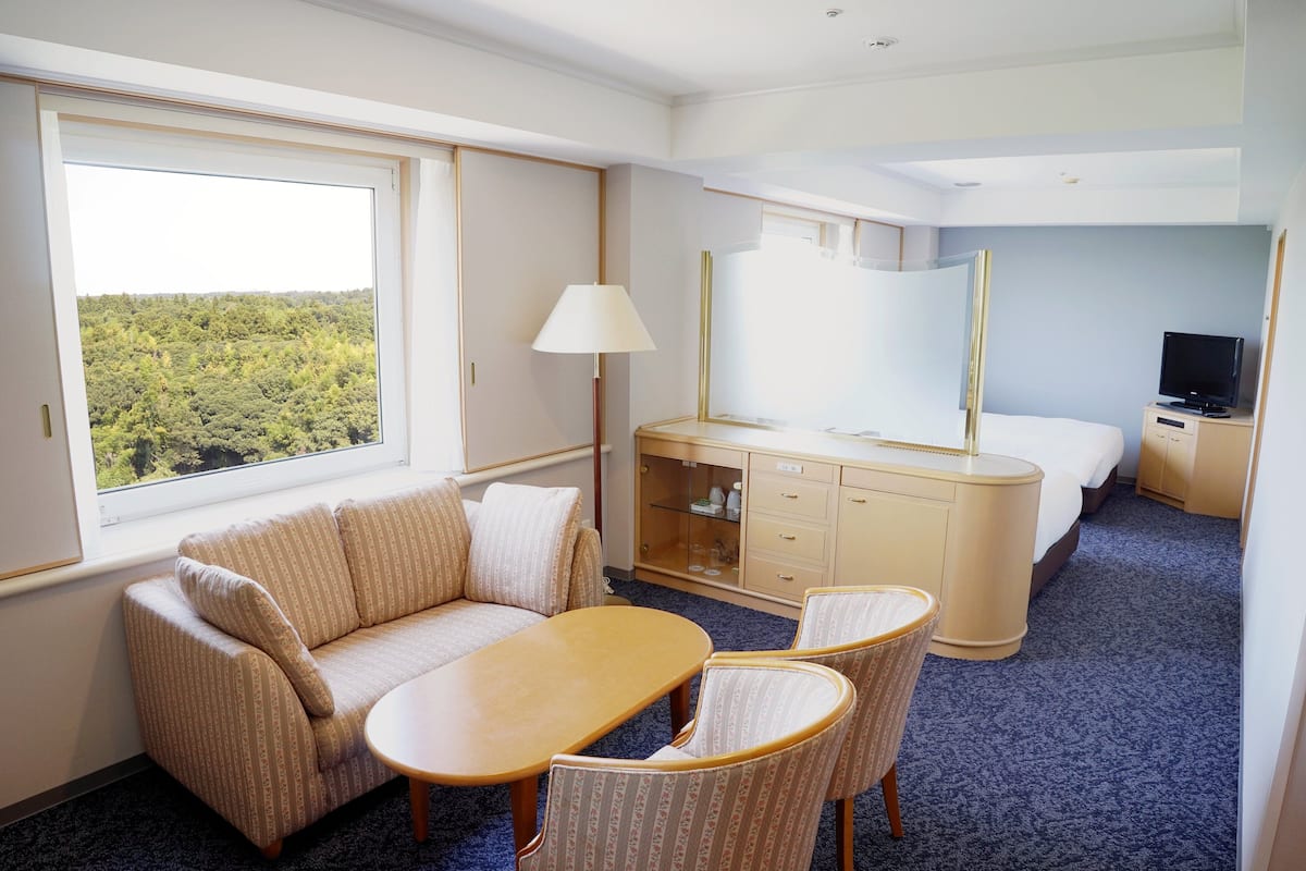 A hotel room with a sofa, a table, a lamp, a bed, a TV, and a dresser.