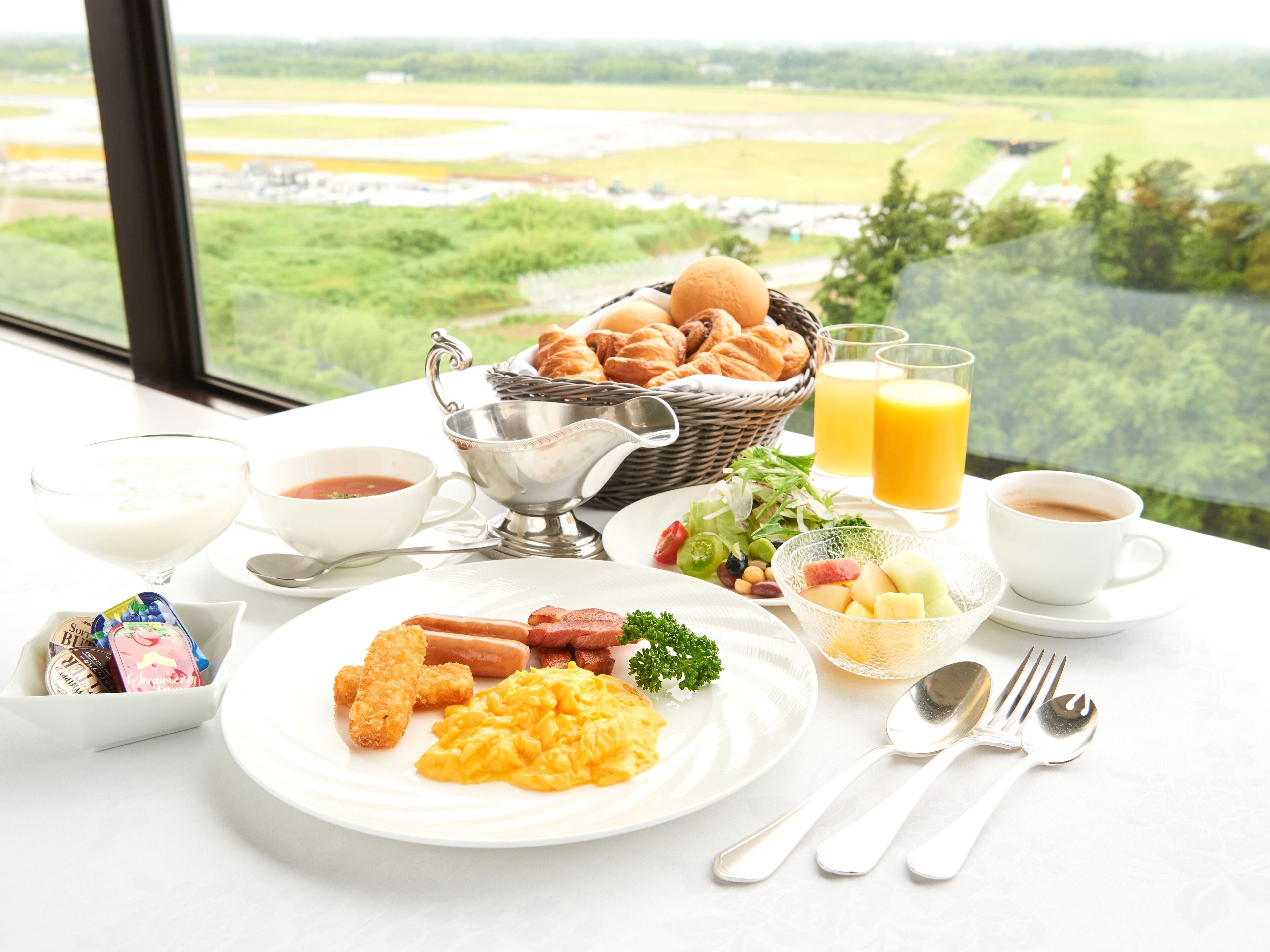 A breakfast table with croissants, scrambled eggs, sausages, and a fruit bowl.