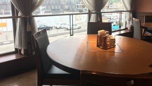 A round table with chairs, a window with curtains, and a view of a street with cars.