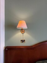 Una lámpara de pared con pantalla de tela, un interruptor rectangular pequeño debajo y un cabecero de madera.